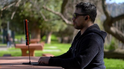 A man in glasses and a black sweatshirt sits in front of an orange and black computer screen just below eye level at the table in front of him. His keyboard sits on the table below. He appears to be in a park as there are trees and grass in the background.