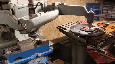 A milling machine with an attached pantograph following the various intricate patterns of a spirograph on the bench next to it. The spirograph is a series of acrylic gears and brass connecting bars mounted on a wooden base.