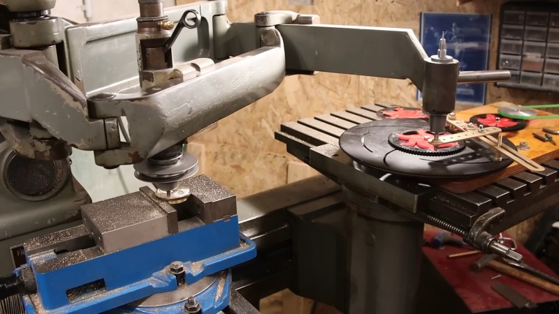 A milling machine with an attached pantograph following the various intricate patterns of a spirograph on the bench next to it. The spirograph is a series of acrylic gears and brass connecting bars mounted on a wooden base.