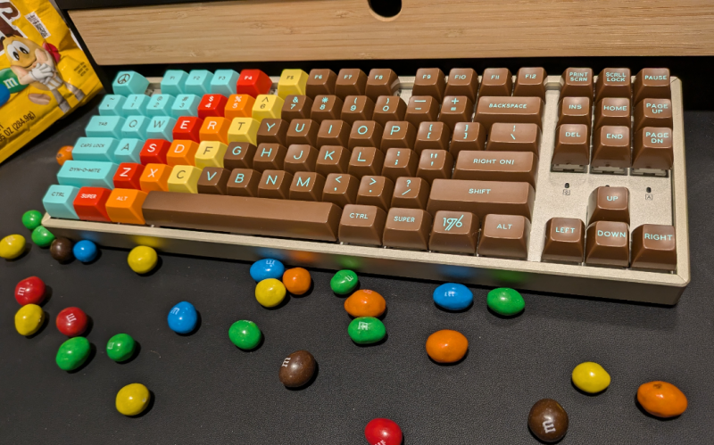 A keyboard with Signature Plastics' 1976 keycap set, with peanut M&Ms scattered around in front of it.