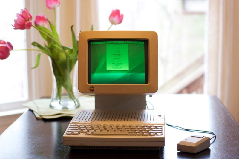 A beige computer monitor with a green glow sits atop a flat, beige Apple IIc with a mouse next to it on a dark wooden table. A vase full of bright pink flowers is in the background.