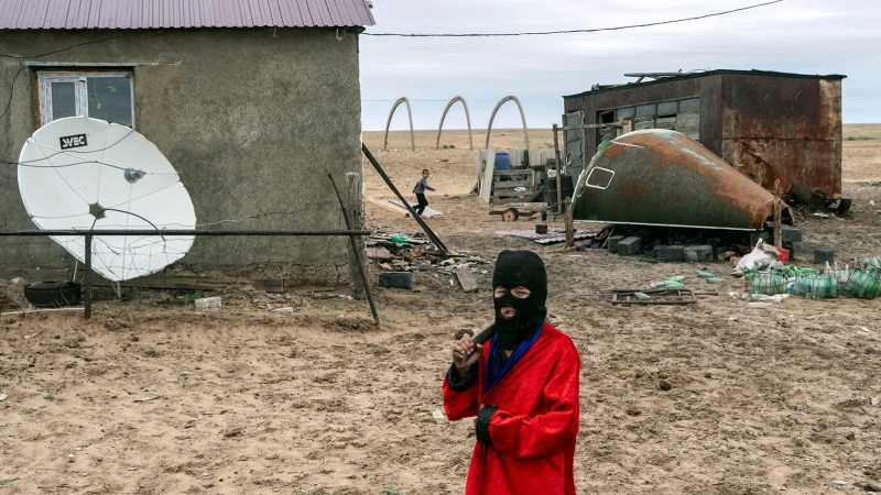 A photo of a farmer in Kazakhstan wearing a balaclava mask standing in front of a farm house with a rusting piece of Soyuz space capsule used as part of the farm's animal feed trough