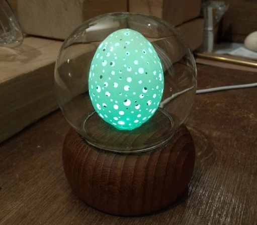 A chicken's egg with many holes glows beneath a glass globe set atop an inverted wooden bowl.