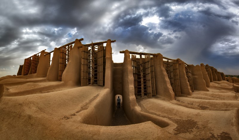 An image of a desert with dramatically cloudy skies. In the middle of the image is a series of clay doorways with vertically-oriented wooden slats surrounding a central pole. These form the basis of a panemone windmill.