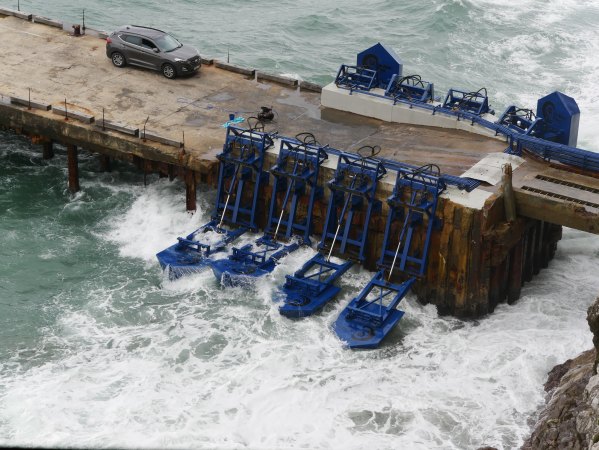 Waves crash near a rocky shore. Large, SUV-sized blue "floaters" sit in the water perpendicular to a concrete pier. The floaters look somewhat like a bass boat shrink wrapped in dark blue plastic and attached to a large piston and hinge. A grey SUV sits on the pier, almost as if for scale.