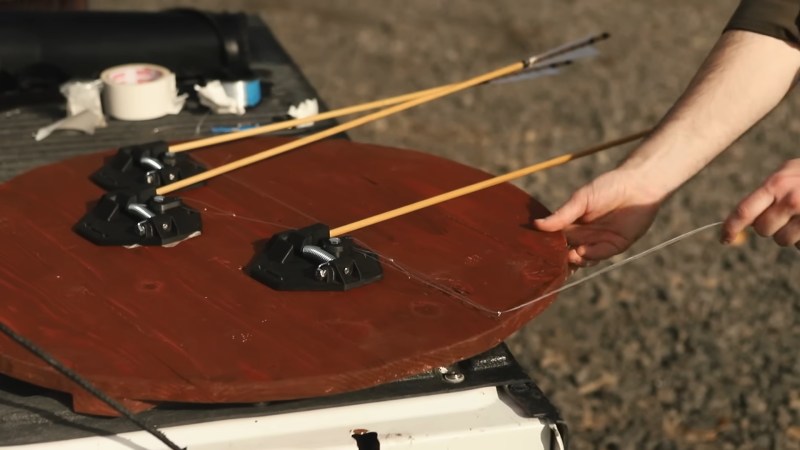 Three 3D printed, spring loaded contraptions sit on a wooden shield. There are arrow shafts connected to the end and a piece of monofilament fishing line extending away from them and through a small eyelet at the edge of. the shield.