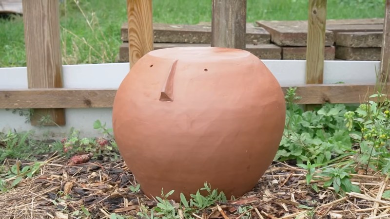 A brown sphere with a flat top, a nose and circular eyes sits on the ground surrounded by low vegetation. A wooden fence is behind it.