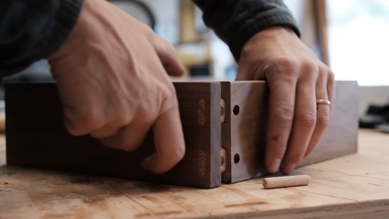 A pair of hands hold two dark brown boards perpendicular two each other on a light brown benchtop. There are two light brown oval dowels in the end of one board that then project toward holes in the opposite board. Circular holes in the oval dowels are visible perpendicular to the second board, and will match up with holes in the board once pressed in. A cylindrical dowel is laying next to the joint and will be placed into the circular holes once assembled.