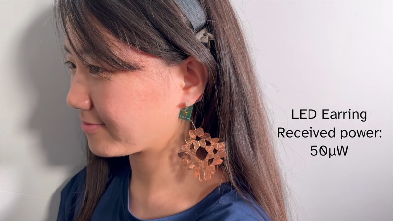 A side view of an Asian woman with brown hair. She has a faint smile and is wearing an earring that looks somewhat like a large copper snowflake. Near the ear hole is a small PCB with a blinking LED. To the right of the image is the text "LED Earring, Recieved power 50 µW"