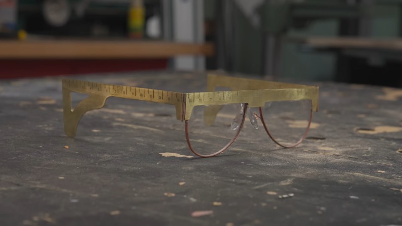 A set of brass safety glasses sit on a marred black workbench. The top and earpiece sections of the frames are in squarish brass plate and have ruler marks on them.