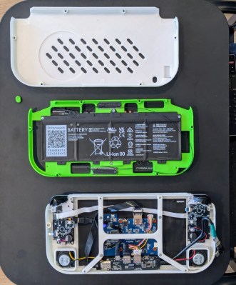 Three 3D printed panels sit on a black surface. The white back has cooling holes in it (top), a green center plate holds a Framework laptop battery (middle), and the front frame holds the speakers, controllers, and screen (bottom)