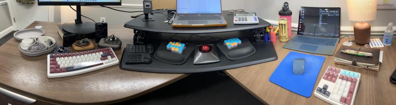 A panorama of OP's desk, featuring a couple of mechanical keyboards and a trackball. 