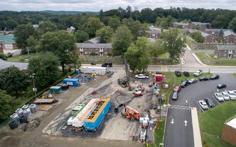 An overhead shot of a parking lot. A road with cars parked along it is on the right hand side of the image. The top center shows a drilling rig on tracks drilling at a slight angle into the ground. Many different semi trailers dot the parking along with several different pallets of construction supplies. An excavator and skylift/forklift are also at work in the lot.