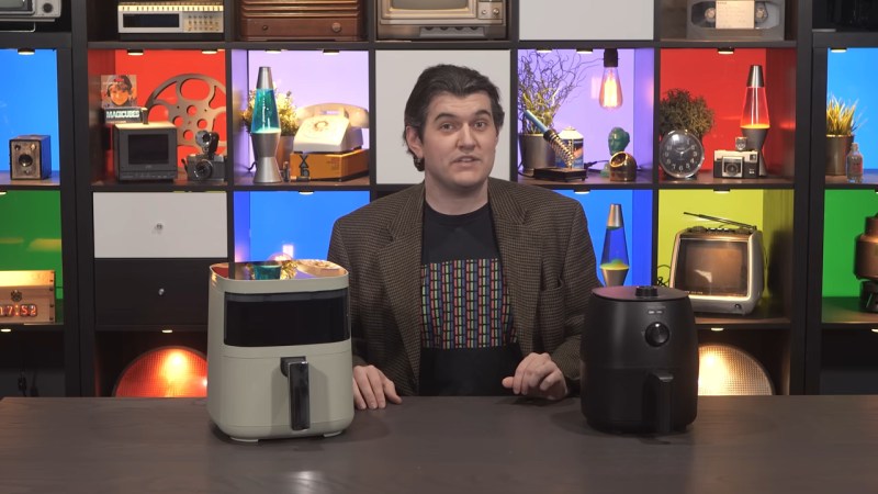 A man in a t-shirt and sport coat sits behind a table upon which are a small, black air fryer and a larger pale green model that looks more futuristic. Behind him is a black set of cube shelves (possibly IKEA Kallax) with different colored interiors lit and holding various bits of vintage technology equipment like landline phones and HiFi setups.