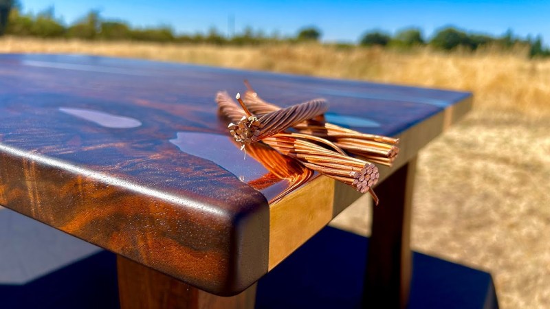 A dark walnut table sits in the sun in what appears to be a field. Voids in the natural wood slab have been filled with shiny bronze, and a bundle of copper wire sits upon the edge of the table in a jaunty artistic fashion.