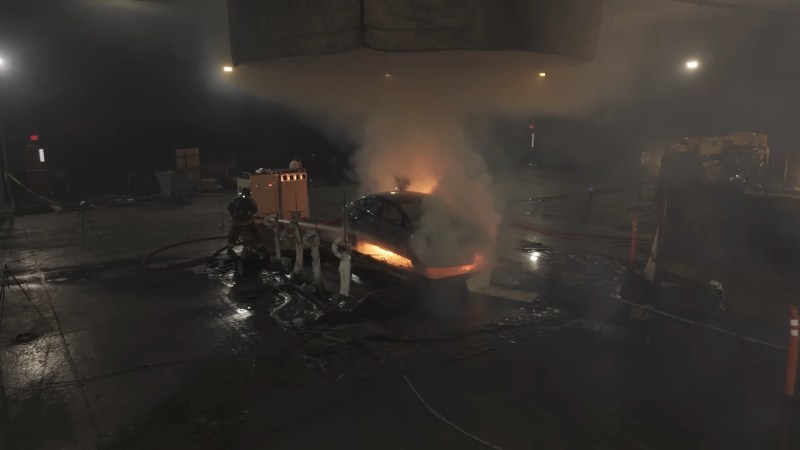 A dark warehouse contains a number of large blocky objects. A Tesla Model 3 sedan sits in the center with flames underneath and curling up the side away from the camera. A firefighter on the left side attempts to put out the fire with a fire hose.