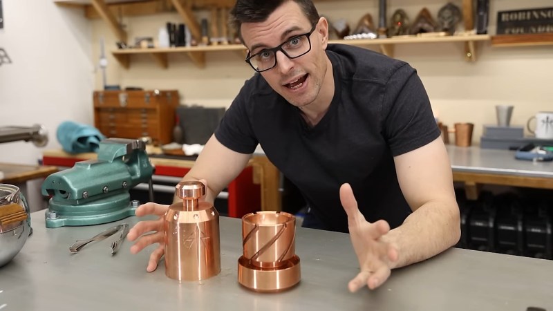 A man in black glasses and a black t-shirt has his arms resting on a grey workbench. Between his opened hands are the two halves of a copper ice press. They are fist-sized copper cylinders. The lower half has large spiraling grooves to aid in the release of excess water from the ice being formed as it melts.