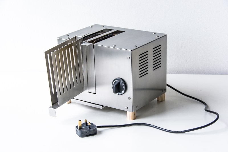 A stainless steel metal toaster sits on a white table. Its cord is draped artfully around to the front and the leftmost toast holding apparatus is rotated out from the front of the device like a book pulled down and out from a bookshelf.