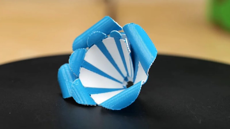 A blue and white, 3D printed rose sits on a black surface with a fuzzy tan background behind it.