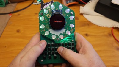 A pair of hands holds the 2024 EMF Tildagon badge with a QWERTY keyboard Hexpansion built by [EastMakes].