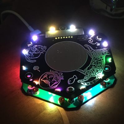 A hexagonal printed circuit board event badge on a table top in the dark, illuminated by coloured LEDs around its edge.