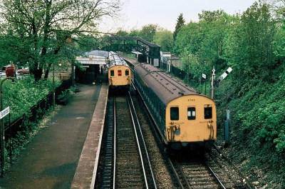 Two 1980s British rail trains with bright yellow ends, in a small British railway station. It's early summer, so the trees surrounding the station are in full leaf.