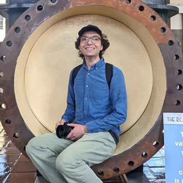 An image of a college student in glasses and a cap sitting inside what appears to be a large pipe with a flange with bolt holes. He is holding a black camera in his lap.