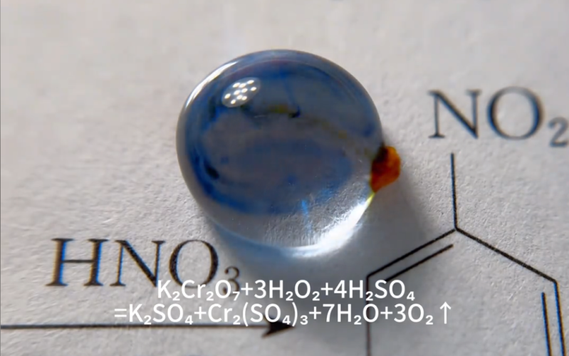 A picture of a single water droplet on top of what appears to be a page from a chemistry text. An orange particle is attached to the right side of the droplet and blue and black tendrils diffuse through the drop from it. Under the water drop, the caption tells us the reaction we're seeing is 