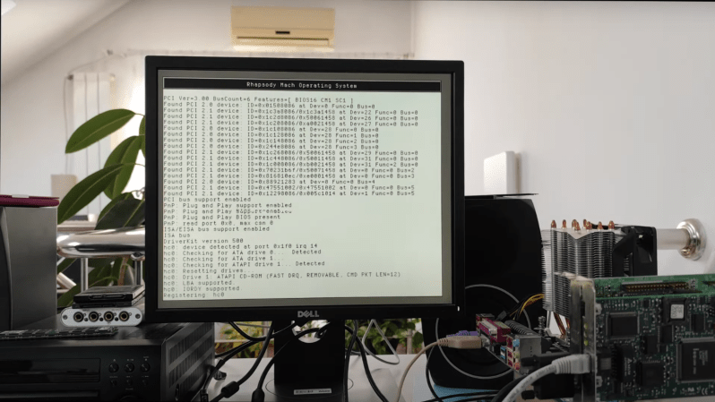 Rhapsody OS is shown in its boot sequence on a monitor; the edge of the motherboard running it is just visible in the right side of the image.