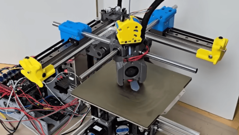 A 3D printer is in the process of printing a test piece. The printer has two horizontal linear rails at right angles to each other, with cylindrical metal rods mounted horizontally on the rails, so that the rods cross over the print bed. The print head slides along these rods.
