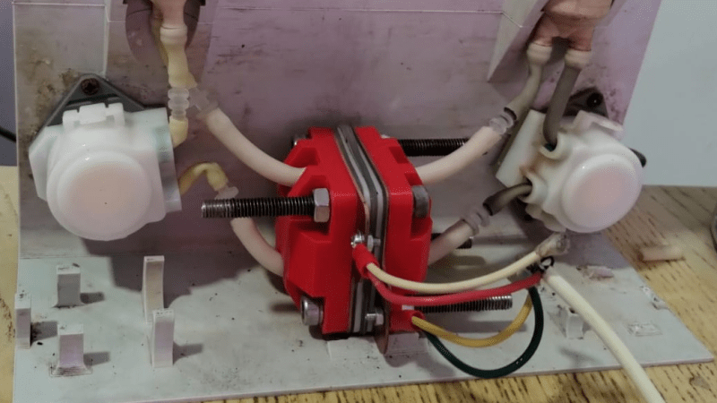 A red, cuboid electrochemical cell is in the center of the picture, with a few wires protruding from the front. Tubes run from each side of the cell to a peristaltic pump and tank on each side. The frame holding the pumps and tanks is white 3D printed plastic.