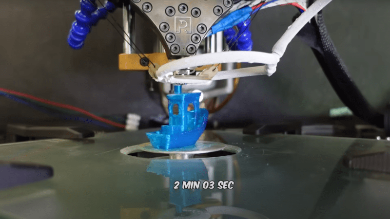 A blue 3DBenchy is visible on a small circular plate extending up through a cutout in a flat, reflective surface. Above the Benchy is a roughly triangular metal 3D printer extruder, with a frost-covered ring around the nozzle. A label below the Benchy reads “2 MIN 03 SEC.”