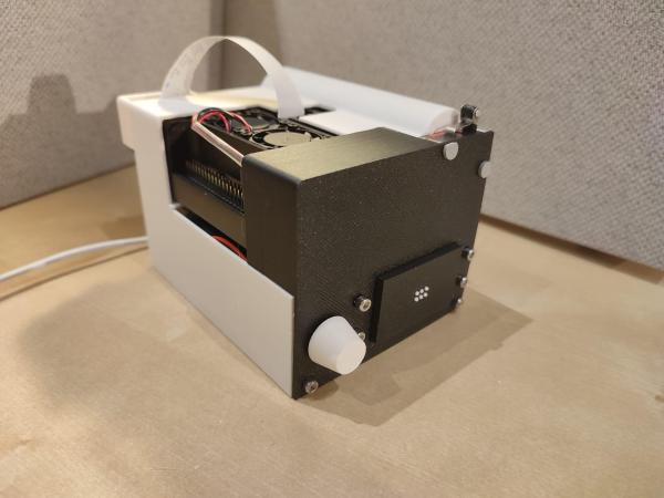 A black and white device sits on a beige table. A white rotary knob projects out near the base of it's rectangular shape nearest the camera. Near it is a black rectangular section of the enclosure with six white dots protruding through holes to form a braille display. A ribbon cable snakes out of the top of the enclosure and over the furthest edge of the device, presumably connecting to a camera on the other side of the device.