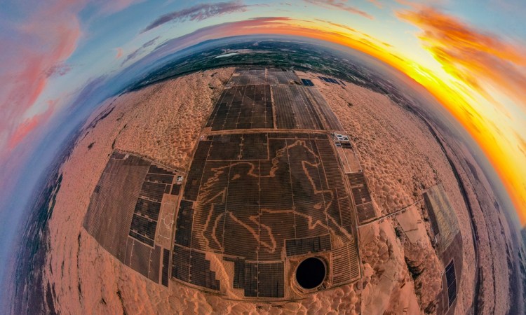 A fisheye lens picture over the Junma Solar Power station in the Mongolian desert. There is a large image of a horse made out of solar panels in the image. A sunset is visible in the upper right of the image, but most the picture is brown sand where there aren't dark blue solar panels.