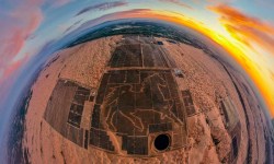 A fisheye lens picture over the Junma Solar Power station in the Mongolian desert. There is a large image of a horse made out of solar panels in the image. A sunset is visible in the upper right of the image, but most the picture is brown sand where there aren't dark blue solar panels.