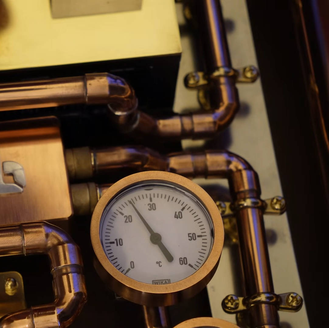 The image shows a close-up view of a temperature gauge mounted among a network of copper pipes. The gauge has a circular face with a white background and black markings, measuring temperature from 0 to 60 degrees Celsius. The needle on the gauge points slightly below 30 degrees Celsius. The copper pipes around the gauge are connected with various fittings and secured with metal brackets, creating an intricate, industrial appearance.