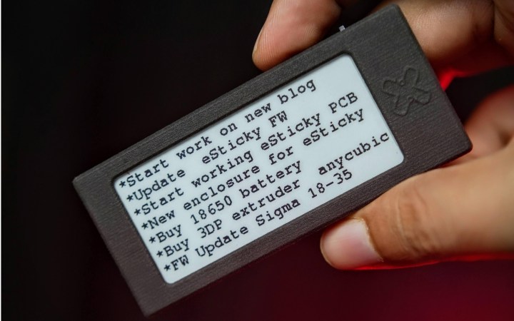 A hand holds a charcoal-colored rectangle with a black and white screen in taking up most of its face. A bulleted list of items are displayed: "Start work on new blog, Update eSticky FW, Start working on eSticky PCB, New enclosure for eSticky, Buy 18650 battery, Buy 3DP extruder anycubic, FW Update Sigma 18-35."