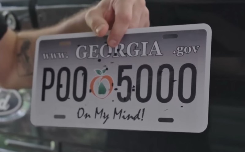 A man holds a license plate in front of a black pickup (F-150 Lightning) tailgate. It is a novelty Georgia plate with the designation P00-5000. There are specks of black superimposed over the plate with a transparent sticker, giving it the appearance of digital mud in black.
