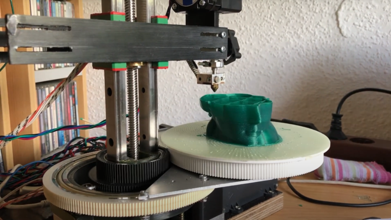 Two geared plastic disks can be seen on a platform. One disk rotates around a central column, while the other is mounted on a platform that extends from the edge of the first disk. The second disk holds a print bed, and a print head mounted on the column is positioned just above a half-finished 3D print.