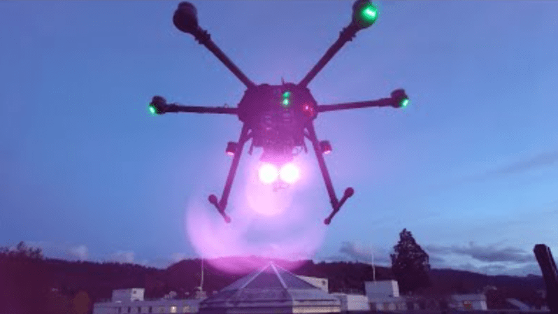 A drone is shown hovering in the sky, with two bright lights shining from its underside.