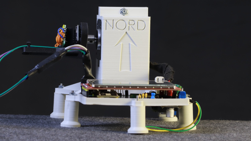 A 3D-printed assembly standing on short legs is visible. A portion extends upward with the word "Nord" sunk into it. Cables extend from one side of the upright portion, and a side view of a circuit board is visible at the front of the assembly.