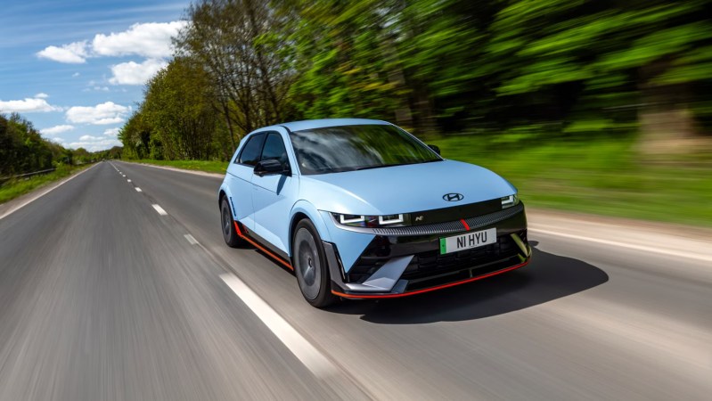 A baby blue hatchback with red accents drives down a road with blurry trees and a blue sky in the background.
