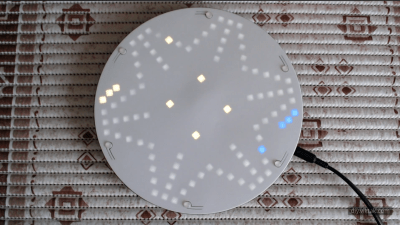 A circular 3D-printed board is shown, with a roughly star-shaped pattern of white LEDs glowing through the surface. Yellow and green LEDs are also visible through the surface at a few points.