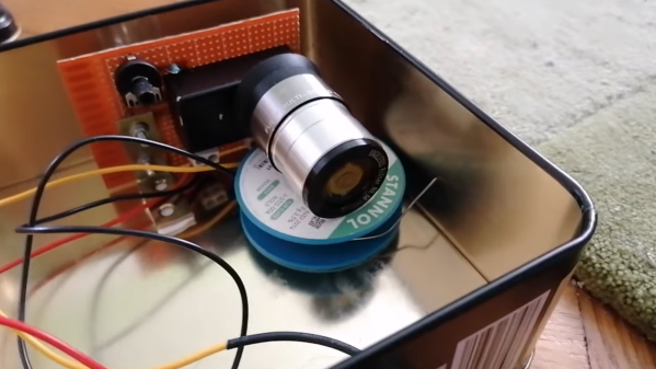A microscope objective is sitting on a spool of solder in a metal tin, in front of a circuit board which has wires running away from it.