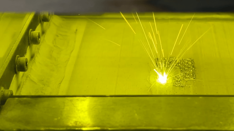 A bed of metal powder is visible through a green-tinted window. A fused metal pattern, roughly square, is visible, with one corner glowing white and throwing up sparks.