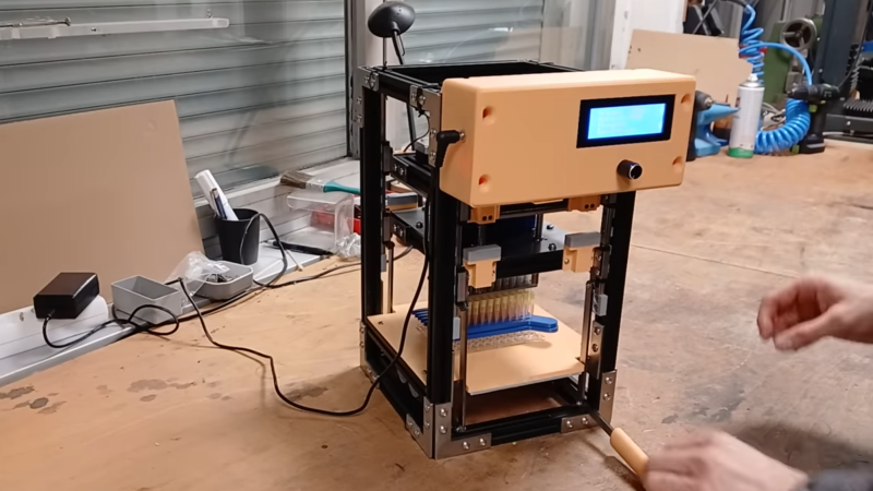 A device within a vertical rectangular frame is shown, with a control box on the front and an LCD display. Within the frame, a grid of syringes is seen held upright beneath two parallel plates.