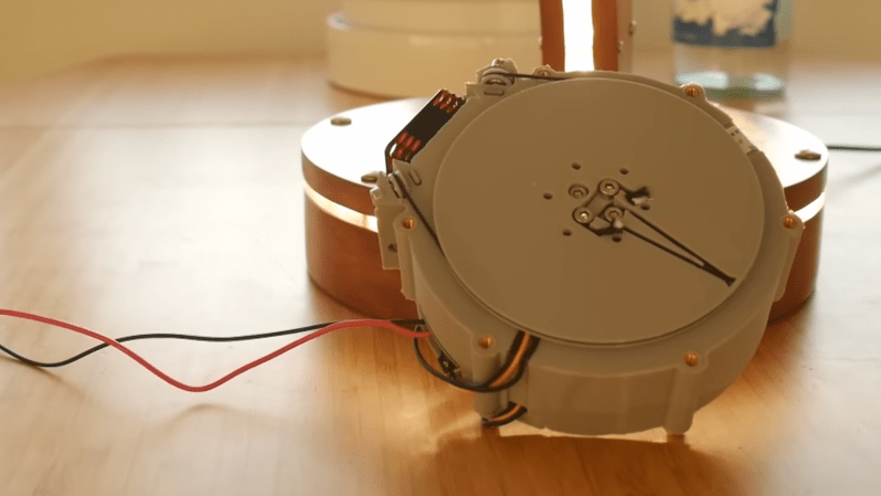 A round, 3D-printed motor housing is shown, with one flattened side holding a fan mount. A circular plate is mounted above the face of the housing, and a cord runs around it and pulleys on the side of the housing.