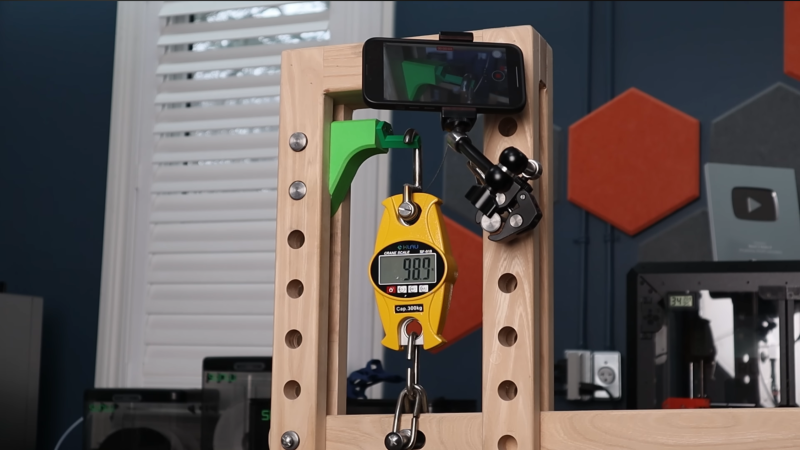 A wooden frame is shown with a scale pulling down on a 3D-printed part held in the frame. A phone on a stand is taking video of the part.
