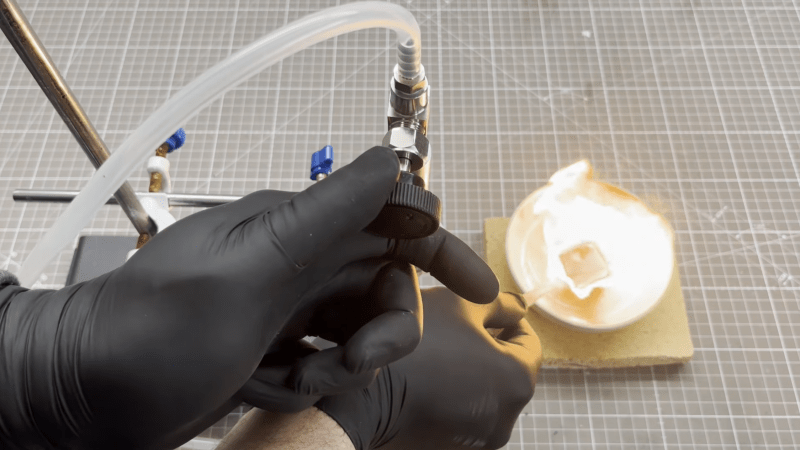 A man's gloved hand is need adjusting the valve on a cylinder, from which a clear plastic tube extends. The man's other hand is seen holding the the other end of the tube in front of a dish of burning wax, which is flaring brightly.