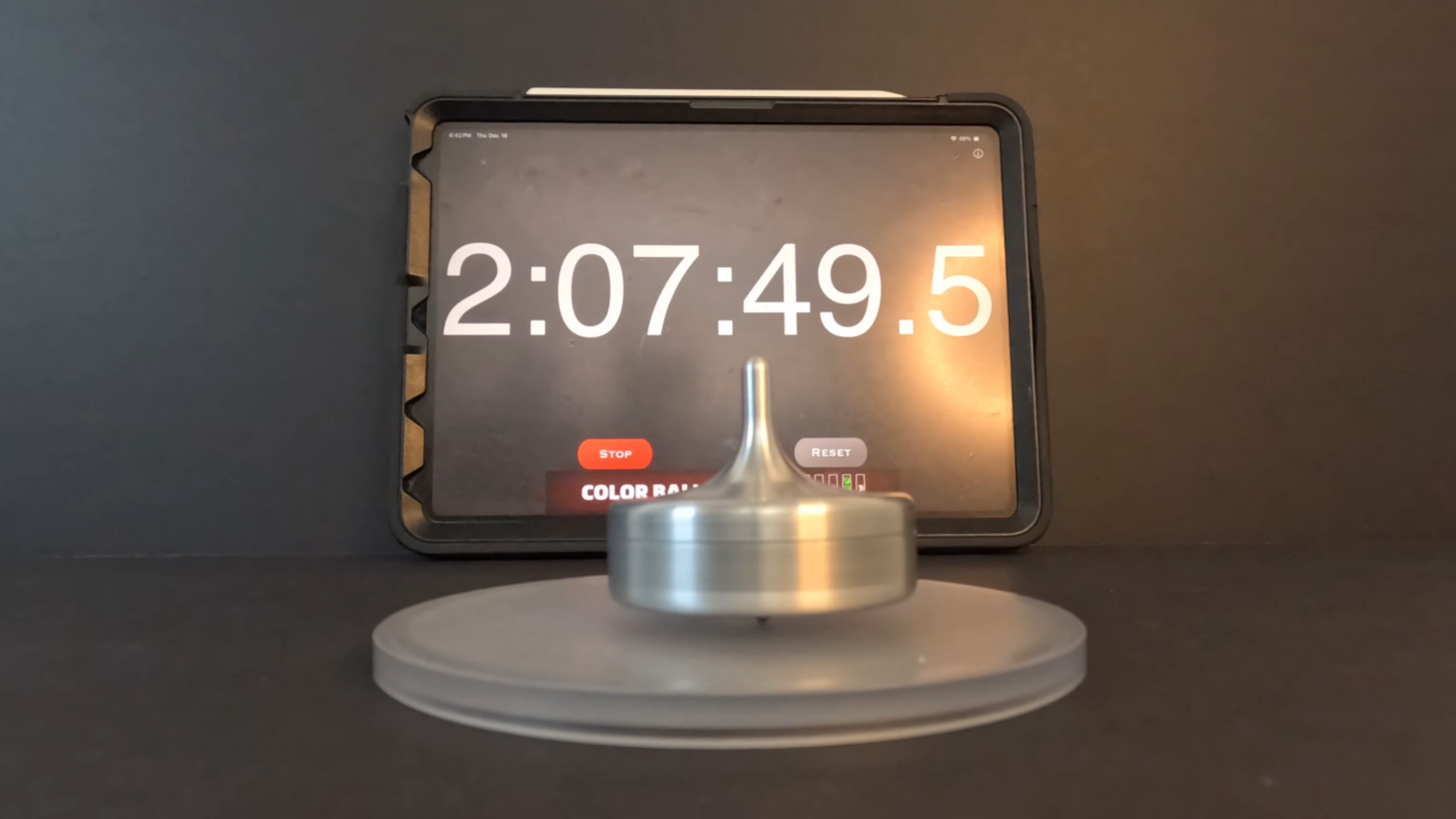 An aluminium top is shown spinning on a plastic disk in front of a tablet showing the text "2:07:49.5"
